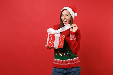 欢快的年轻圣诞老人女孩穿着针织毛衣圣诞帽，手里拿着打开的红色盒子，礼物孤立在红色背景上。 新年快乐，2019年庆祝节日聚会的理念