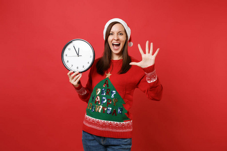 开朗的年轻圣诞老人女孩拿着圆钟，展示手掌张开的手指孤立在红色的背景上。 时间不多了。 新年快乐，2019年庆祝节日聚会的理念。 