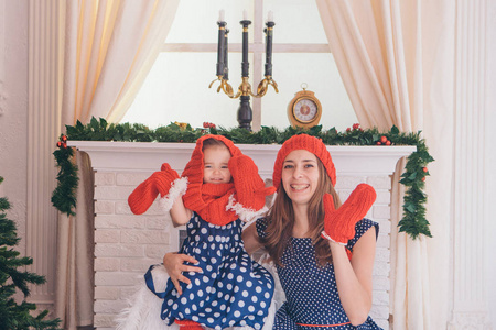 一个女人带着一个孩子，穿着蓝色的裙子，红色的帽子上有圆点，里面有手套，背景是圣诞节壁炉，圣诞树，礼物，手表