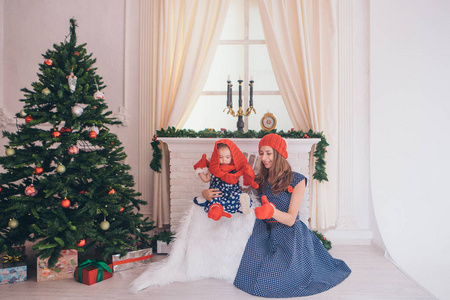 一个女人带着一个穿着蓝色连衣裙的孩子，戴着红色帽子和手套的圆点，背景是圣诞节内部壁炉圣诞树礼物手表