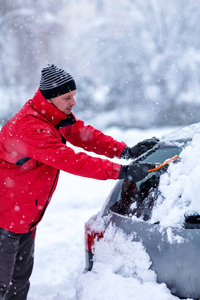 年轻人把雪从车里移走。 男人用刷子把雪从汽车挡风玻璃上清理出来。 下雪的冬天天气。 暴风雪后雪中的汽车。