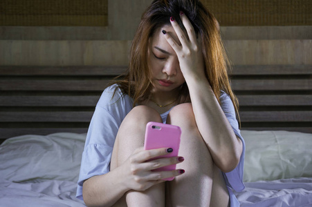 年轻抑郁的亚洲华裔女孩晚上在床上用手机哭泣，感到悲伤和沮丧，网络欺凌或心碎的受害者，绝望的女人的表情抑郁