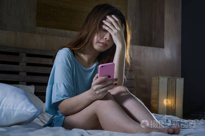 年轻抑郁的亚裔中国女孩晚上用手机在床上哭泣,感到悲伤和沮丧,网络