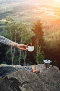 女孩手拿着一杯热咖啡靠近篝火，背景是自然在日落。 概念冒险活动户外度假。 夏令营。