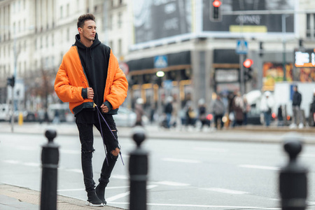 时髦的在建筑物附近的街道上。 穿橙色夹克，全是黑色的。 冬季秋装。 夹克衫和黑色运动鞋。