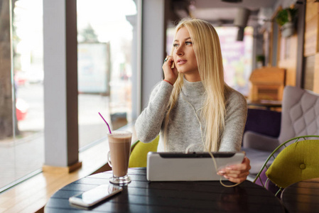 年轻的金发女人用平板电脑喝咖啡。 在舒适的咖啡馆里放松，坐在椅子上，愉快地听着耳机有声书或音乐