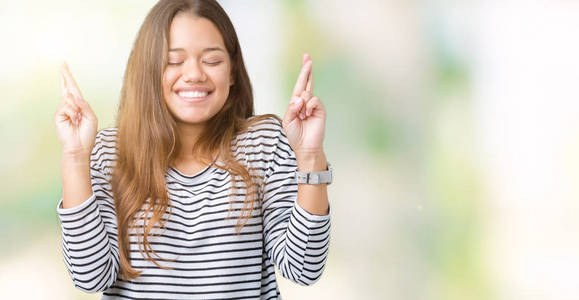 年轻漂亮的黑发女人穿着条纹毛衣，在孤立的背景上微笑，交叉手指，希望和眼睛闭上。 运气和迷信的概念。