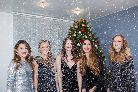 新年节日庆典派对的概念。一群美丽迷人的女人，在圣诞树和五彩纸屑的背景下，化着亮丽的妆容，穿着华丽的礼服