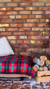 沙发附近的一叠桦木，在敞开的冬季雪覆盖的露台上铺着一座乡间房子的砖墙。 冬天小屋的舒适内部