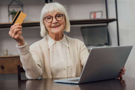 戴眼镜的老妇人坐在电脑桌旁，拿着信用卡，在家里网上购物