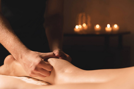 男性手的特写在黑暗的房间里做小腿按摩女性腿在背景蜡烛。美容和水疗护理