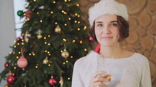 微笑的妇女在圣诞老人帮助帽与一杯香槟酒在客厅和圣诞树背景