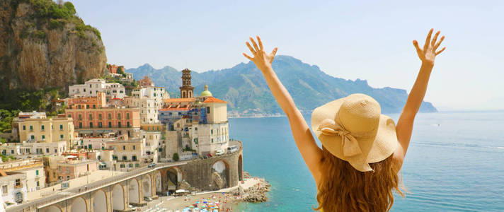 意大利的暑假。 全景后景年轻女子带着草帽，举起双臂，看着意大利阿特拉尼村的阿马尔菲海岸。