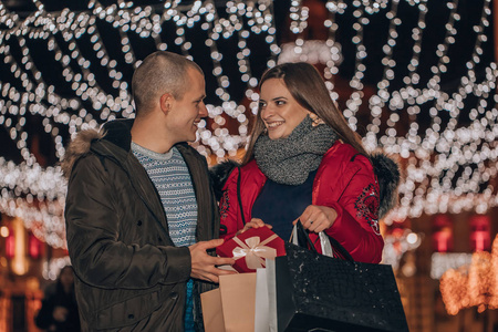 年轻开朗的夫妇拿着购物袋，把礼物放在一个盒子里，在城市的街道上玩得很开心，背景上有很多灯。 图像