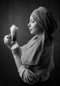 裹着头巾的年轻漂亮女人拿起一杯带奶油的咖啡。 完美的化妆棕色背景。 模型加上尺寸。 黑白的。