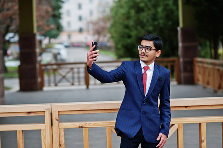 戴眼镜的印度年轻人穿着西装，打着红色领带，在户外摆好姿势，在手机上自拍。