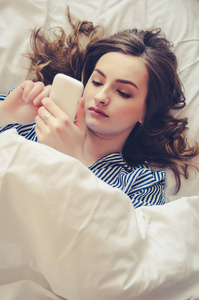 时髦女孩躺在床上时正在手机上阅读男友的短信。 穿睡衣的开朗的年轻女人在睡觉前在手机上看视频。