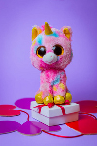 玩具粉红独角兽坐在一个白色礼品盒上，红色丝带在纸心上的紫色背景。