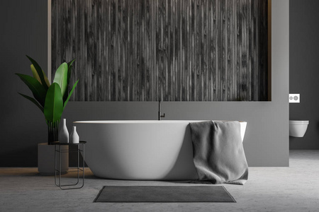 现代浴室内部有灰色和木制墙壁，混凝土地板，白色浴缸，上面有毛巾，地毯靠近它和一个厕所。 3D渲染