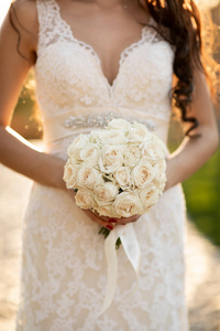 s hands hold a round wedding bouquet of ivory roses. Sunset warm