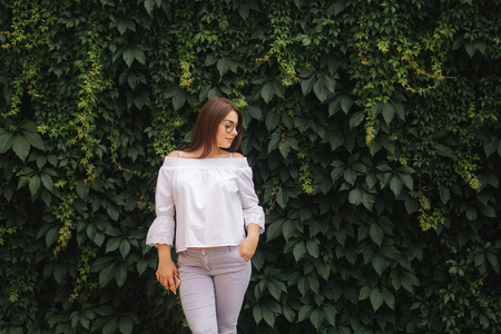 太阳镜中的快乐女性肖像。女孩穿着白色的衬衫站在附近的墙壁杂草丛生常春藤