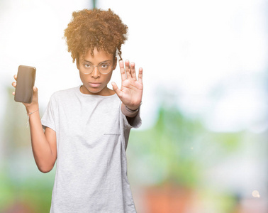 年轻的非洲裔美国妇女显示智能手机屏幕在孤立的背景上，张开手，做停止标志，严肃和自信的表情，防御手势。