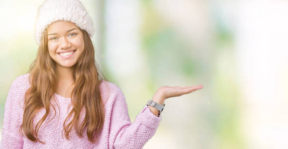 年轻漂亮的黑发女人，穿着毛衣和冬帽，在孤立的背景上微笑，欢快的呈现，用手掌指着相机。