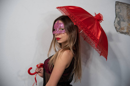 美丽的年轻女子肖像与威尼斯狂欢节红色面具与红色伞。 魅力女孩与完美的化妆金发与透明坦克顶部和粉红色胸罩。 面具派对的迷人照片