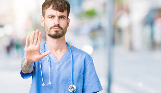 年轻英俊的护士穿着外科医生制服，在孤立的背景下做停止用手掌唱歌。 警告表情，脸上有负面和严肃的手势。