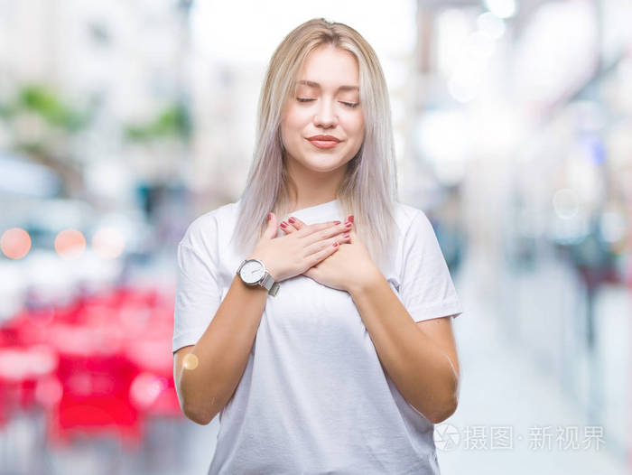 年轻的金发女子在孤立的背景上微笑,双手放在胸部,闭着眼睛,脸上有