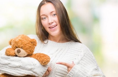 年轻的白种人女人抱着玩具熊在孤立的背景下非常高兴地用手和手指指着