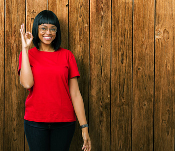 美丽的年轻非洲裔美国妇女戴着眼镜，在孤立的背景上微笑，积极地用手和手指做OK标志。 成功的表达。