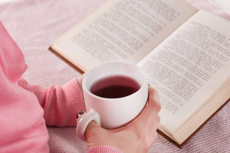 女人在家里的床上拿着一杯热茶饮料和看书。女孩穿粉色羊毛衫。育和寒冷天气理念..把注意力集中起来