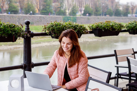 年轻的女学生在公园学习，在阳光明媚的日子里使用现代笔记本电脑，微笑的女孩在户外咖啡馆坐着工作