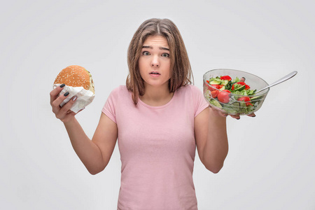 沮丧的年轻女子手里拿着汉堡和沙拉碗。隔离在灰色背景上