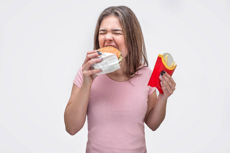 饥饿的年轻女子咬一块汉堡。她用另一只手拿着炸土豆。隔离在白色背景上