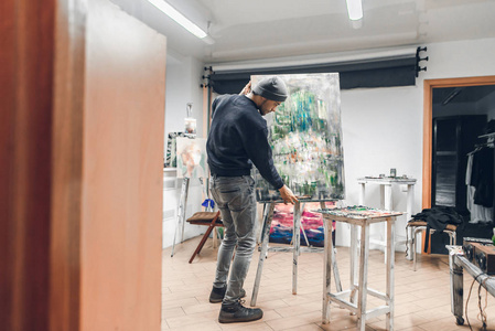 艺术家穿着一件深色毛衣和帽子在工作室工作，一个人把一幅未完成的图片放进画架，并将继续画。 艺术家在一个舒适的艺术工作室的肖像。 