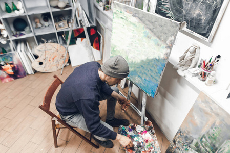 有创意的人坐在一个工作室的椅子上，靠近一个干净的主人，在调色板上混合一幅画，画一幅新画。 上面的风景。 在工作室的家里创作一幅油