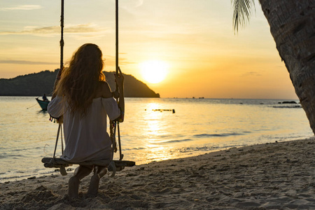 一个女孩的剪影，她坐在美丽的黄昏海滩上的普吉岛上的秋千上，寻找日落海滩和山