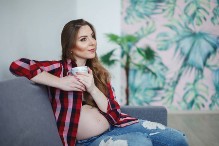 一位身穿格子衬衫长发的美丽孕妇坐在灰色沙发上，的肚子手里拿着一杯茶。 幸福孕产健康生活方式的概念。