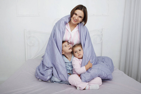 年轻漂亮的母亲坐在毯子下，孩子们躺在床上，穿着蓝色的亚麻布。