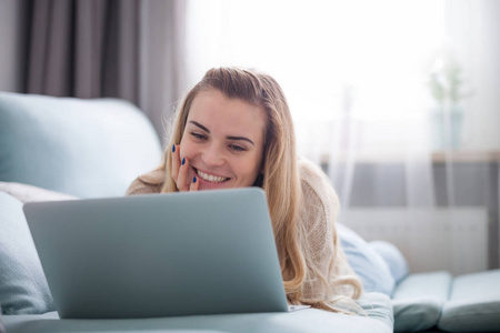 微笑的女人躺在家里的沙发上看着笔记本电脑屏幕，放松和休息