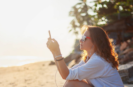 美丽的年轻女孩，坐在海滩上，戴着耳机听音乐休息和度假
