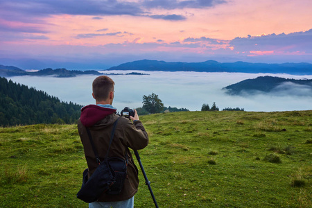 专业摄影师在岩石峰上用相机拍摄照片。 如梦似幻的雾中风景春天橘红色的雾中日出在下面美丽的山谷