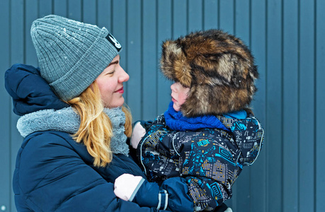 年轻快乐的女人，她的儿子抱着她的蓝色灰色栅栏背景在冬天。 冬帽