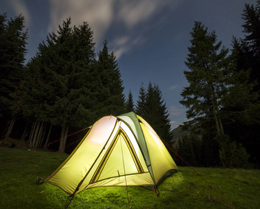 旅游徒步旅行者的帐篷从里面明亮地照亮了绿色的草地森林，在遥远的山脉背景下，深蓝色的星空下，高大的松树之间空地。 晚上在山上露营。