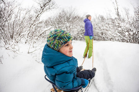 一个女人把一个男孩卷在雪橇上。 一个孩子和他的母亲一起穿过冬天的森林。 脸颊红润的孩子坐着雪橇在雪地里骑着。 寒假。 和父母一起