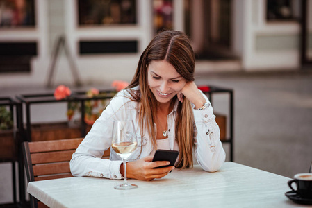快乐的年轻女人坐在户外餐厅看智能手机。 桌子上有一杯白葡萄酒。