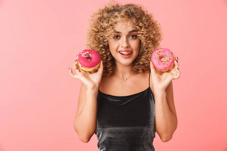 开朗的年轻女人，卷发，孤立地站在粉红色的背景上，吃着釉面圈