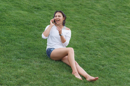 快乐美丽的年轻女子坐在草地上，用智能手机